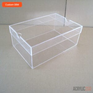 Clear acrylic shoe box on wooden table under bright light — semicircle finger-notch handle on lid visible, high-transparency PMMA sneaker storage box